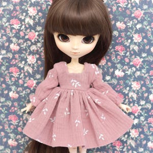 Peut inclure: Une robe de poup&eacute;e rose avec des broderies de feuilles blanches. La robe a des manches longues et une jupe pliss&eacute;e. La poup&eacute;e a les cheveux bruns et porte la robe.
