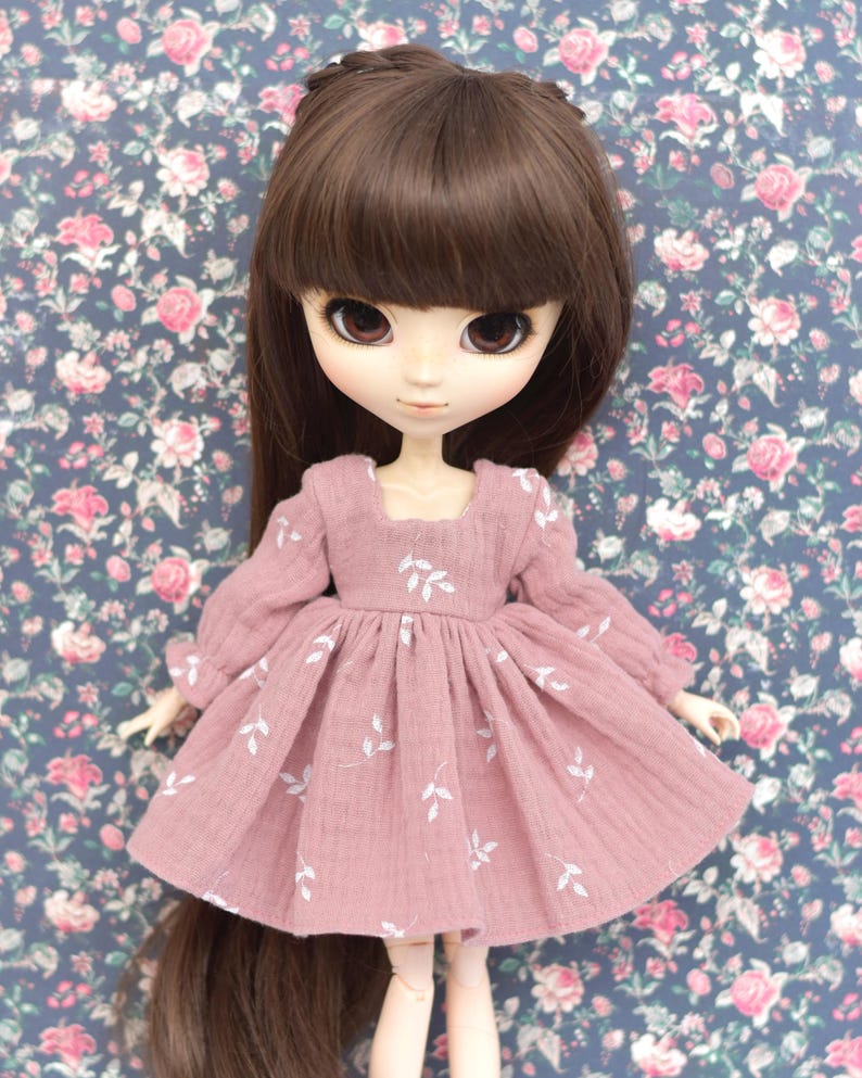 Peut inclure: Une robe de poup&eacute;e rose avec des broderies de feuilles blanches. La robe a des manches longues et une jupe pliss&eacute;e. La poup&eacute;e a les cheveux bruns et porte la robe.
