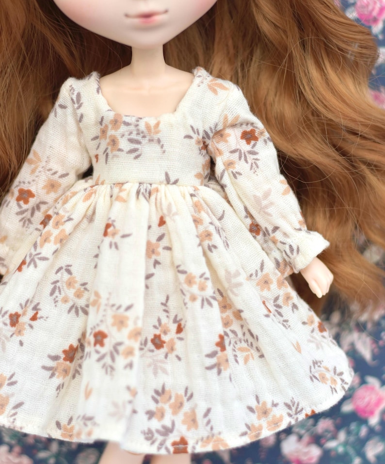 Peut inclure: Une robe blanche &agrave; motif floral dans les tons brun et beige. La robe a des manches longues et une jupe pliss&eacute;e.