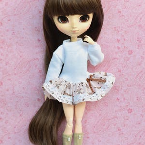 Op de afbeelding: Een pop met lang bruin haar, die een witte trui en een bruin-witte bloemenrok met kant draagt. De pop draagt kniekousen en heeft een roze achtergrond.