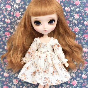 Peut inclure: Une poup&eacute;e avec de longs cheveux ondul&eacute;s auburn portant une robe blanche &agrave; motif floral. La robe a des manches longues et une jupe pliss&eacute;e.