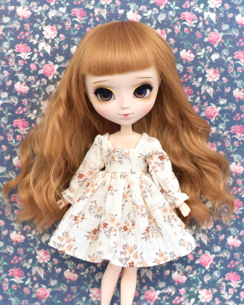 Peut inclure: Une poup&eacute;e avec de longs cheveux ondul&eacute;s auburn portant une robe blanche &agrave; motif floral. La robe a des manches longues et une jupe pliss&eacute;e.
