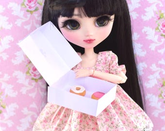 May include: A doll with long black hair wearing a pink floral dress and holding a white box with two donuts inside.