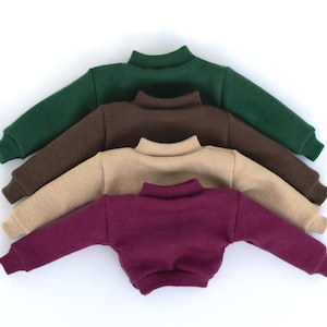 Op de afbeelding: Vier truien gestapeld op elkaar. De truien zijn in verschillende kleuren: groen, bruin, beige en bordeaux.