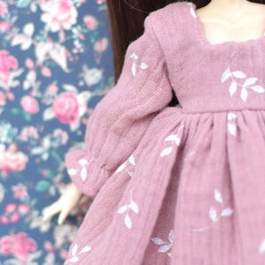 Peut inclure: Une robe rose avec des broderies florales blanches. La robe a un col carr&eacute; et des manches longues.