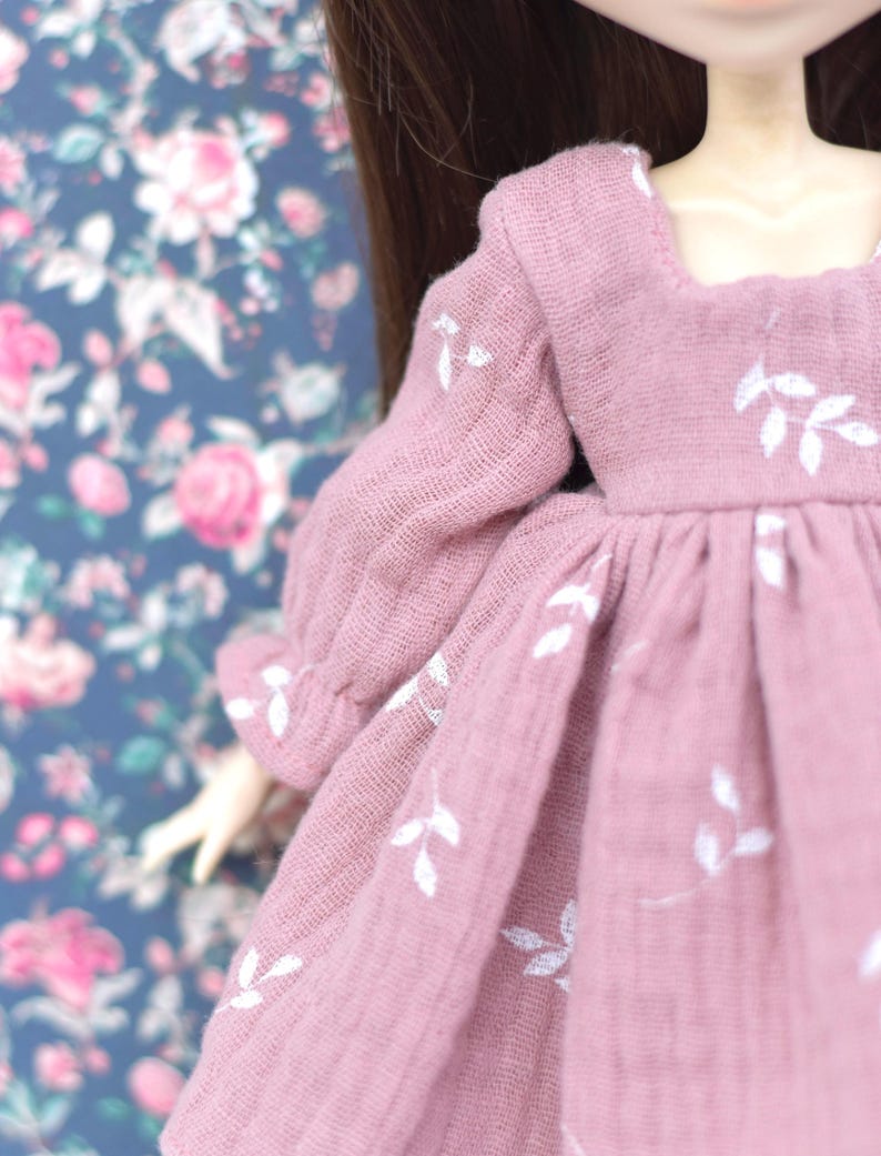 Peut inclure: Une robe rose avec des broderies florales blanches. La robe a un col carr&eacute; et des manches longues.