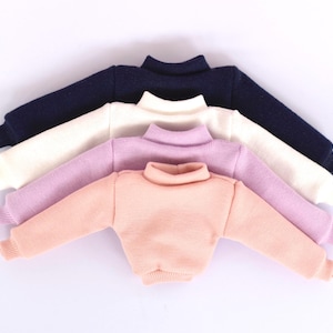 Peut inclure: Quatre petits pulls à col roulé de couleur marine, crème, lavande et pêche. Les pulls sont disposés en rangée, mettant en valeur leurs différentes couleurs et textures. Les pulls ont des manches longues et des poignets côtelés.