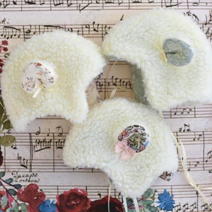 Könnte beinhalten: Drei cremefarbene, flauschige Babyhauben mit floralen Stoffakzenten und Bändern. Die Hauben haben eine abgerundete Form und sind aus einem weichen, strukturierten Material gefertigt. Der Hintergrund zeigt Notenblätter und florale Akzente.