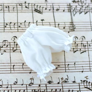 May include: White cotton baby bloomers with a ruffled edge. The bloomers are lying on a sheet of music.