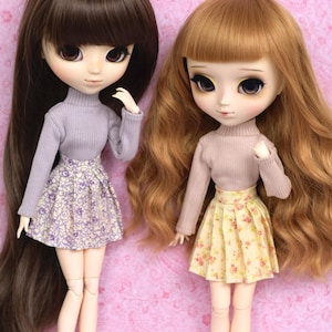 Può includere: Due bambole con lunghi capelli castani e biondi che indossano maglioni abbinati lavanda e rosa e gonne floreali. Le bambole hanno grandi occhi e sono in piedi su uno sfondo rosa.