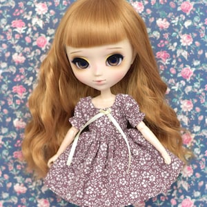 Peut inclure: Une poupée aux longs cheveux roux ondulés et aux grands yeux expressifs. Elle porte une robe violette à motifs floraux blancs et un ruban blanc. Le fond est un motif floral bleu et rose.