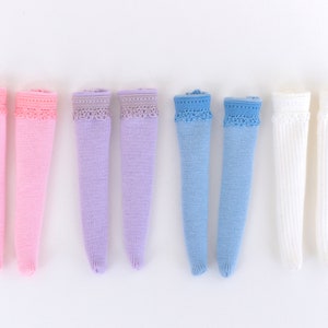 Può includere: Una collezione di calze al ginocchio in vari colori pastello: rosa, lavanda, azzurro e bianco. Ogni calza presenta un delicato bordo in pizzo sulla parte superiore. Le calze sono disposte su uno sfondo bianco.