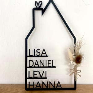 May include: A black, house-shaped wall hanging with the names LISA, DANIEL, LEVI, and HANNA cut out. A small heart tops the chimney. Dried flowers and a twine bow accent the right side. The piece is displayed on a wooden surface.