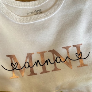 Könnte beinhalten: Ein weißer Baby-Body mit den Worten "MINI" und "hanna" in Roségold-Schrift. Das Wort "hanna" ist mit einer schwarzen Linie und zwei Herzen mit "MINI" verbunden. Der Body von Simple Joys by Carter's liegt auf einer Holzoberfläche.