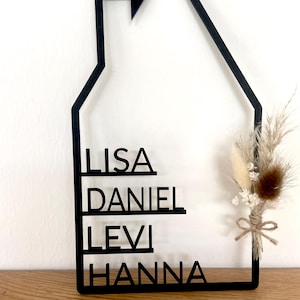 May include: Black wooden house-shaped wall decor with the names LISA, DANIEL, LEVI, and HANNA. A small heart is at the top. A bundle of dried flowers and grasses is tied to the side with twine. The decor is displayed on a wooden surface.