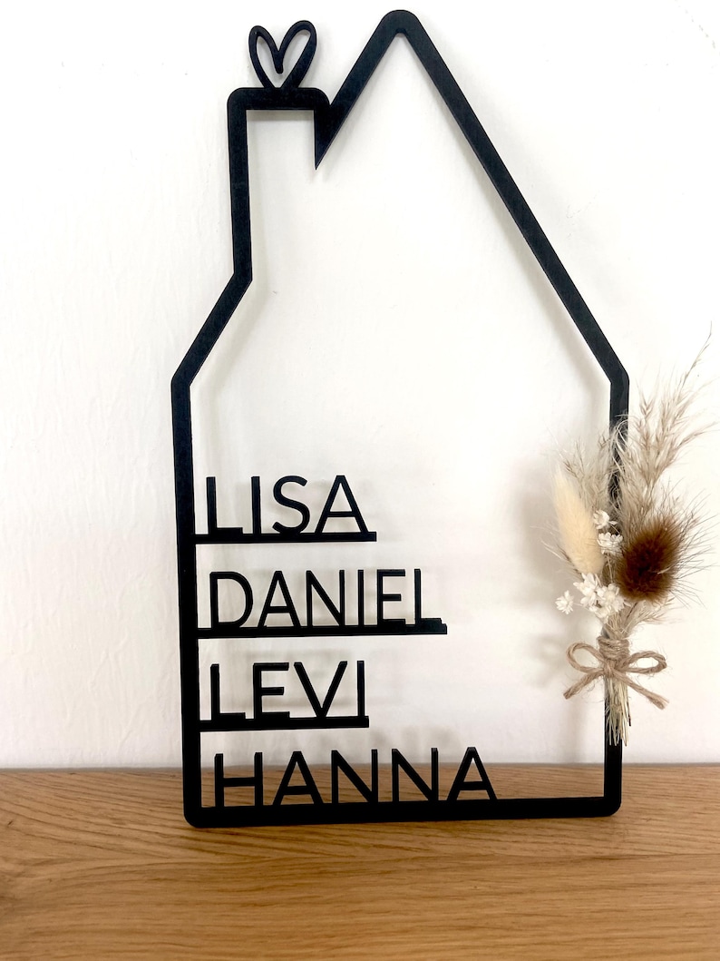 May include: Black wooden house-shaped wall decor with the names LISA, DANIEL, LEVI, and HANNA. A small heart is at the top. A bundle of dried flowers and grasses is tied to the side with twine. The decor is displayed on a wooden surface.