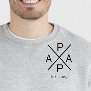 Könnte beinhalten: Ein grauer Pullover mit einem schwarzen Grafikdesign, das die Buchstaben "P A P" in einem durchgestrichenen Design zeigt. Der Text "Est. 2025" befindet sich unterhalb der Grafik.