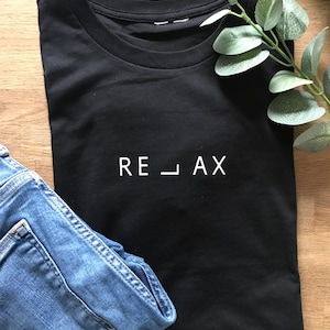 Könnte beinhalten: Schwarzes T-Shirt mit weißem Text, der "RE_LAX" lautet. Das Shirt ist gefaltet und liegt auf einer Holzoberfläche mit einer blauen Jeans und einem Zweig Grün.