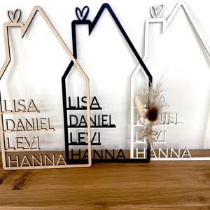 May include: Three wooden house-shaped signs with family names. One is natural wood, one is black, and one is white. Each has a heart detail at the top and includes the names Lisa, Daniel, Levi, and Hanna.