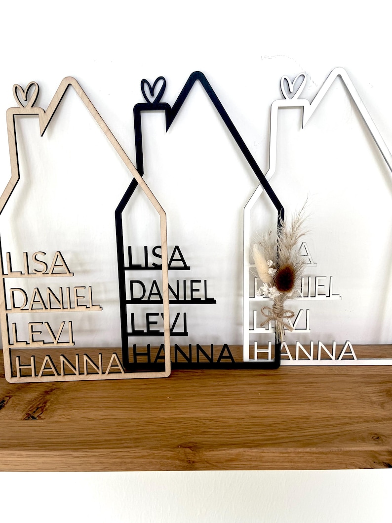 May include: Three wooden house-shaped signs with family names. One is natural wood, one is black, and one is white. Each has a heart detail at the top and includes the names Lisa, Daniel, Levi, and Hanna.
