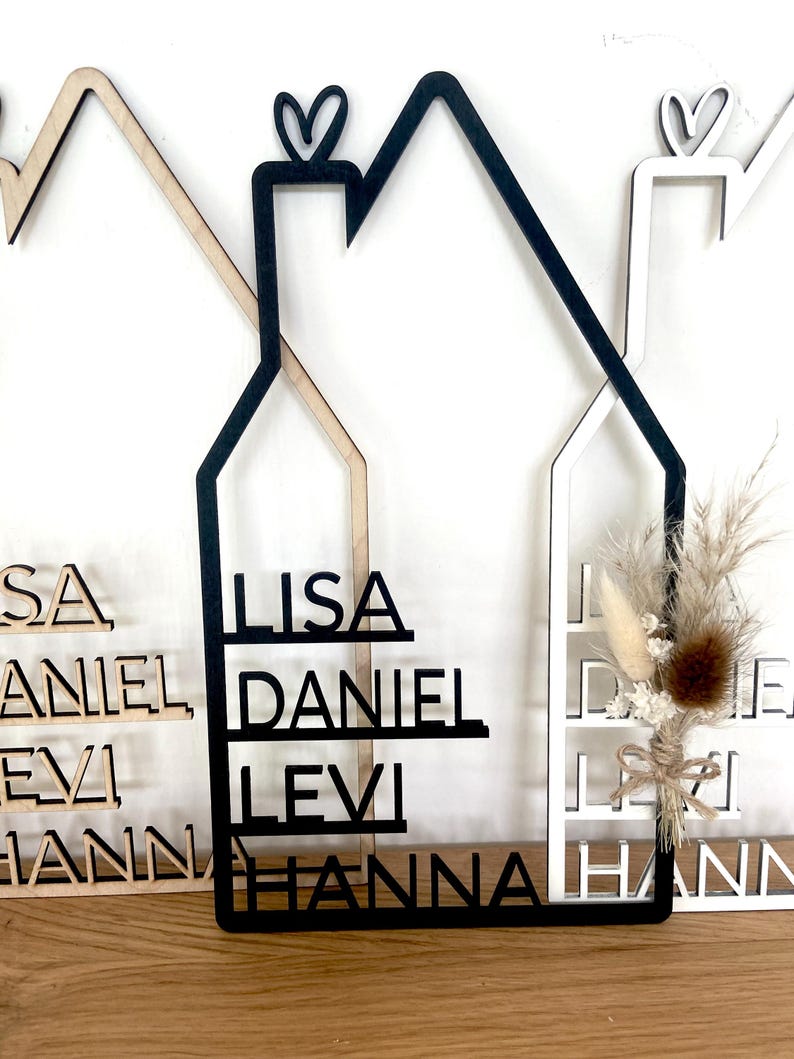 May include: Wooden house-shaped wall decor with the names LISA, DANIEL, LEVI, and HANNA cut out. The house is black with a heart on top. Other houses in white and natural wood are also visible.