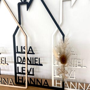 May include: Wooden house-shaped wall decor with family names cut out. The houses are in natural wood, black, and white. One house has a small dried flower arrangement. Each house has a heart shape at the top.