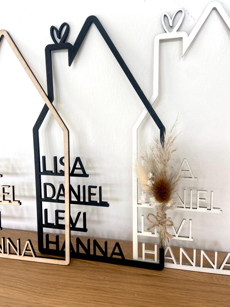 May include: Wooden house-shaped wall decor with family names cut out. The houses are in natural wood, black, and white. One house has a small dried flower arrangement. Each house has a heart shape at the top.