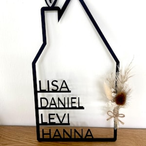 May include: Black wooden house-shaped sign with the names LISA, DANIEL, LEVI, and HANNA. A small heart is at the top. The sign is decorated with dried flowers and tied with twine. The sign is displayed on a wooden surface.