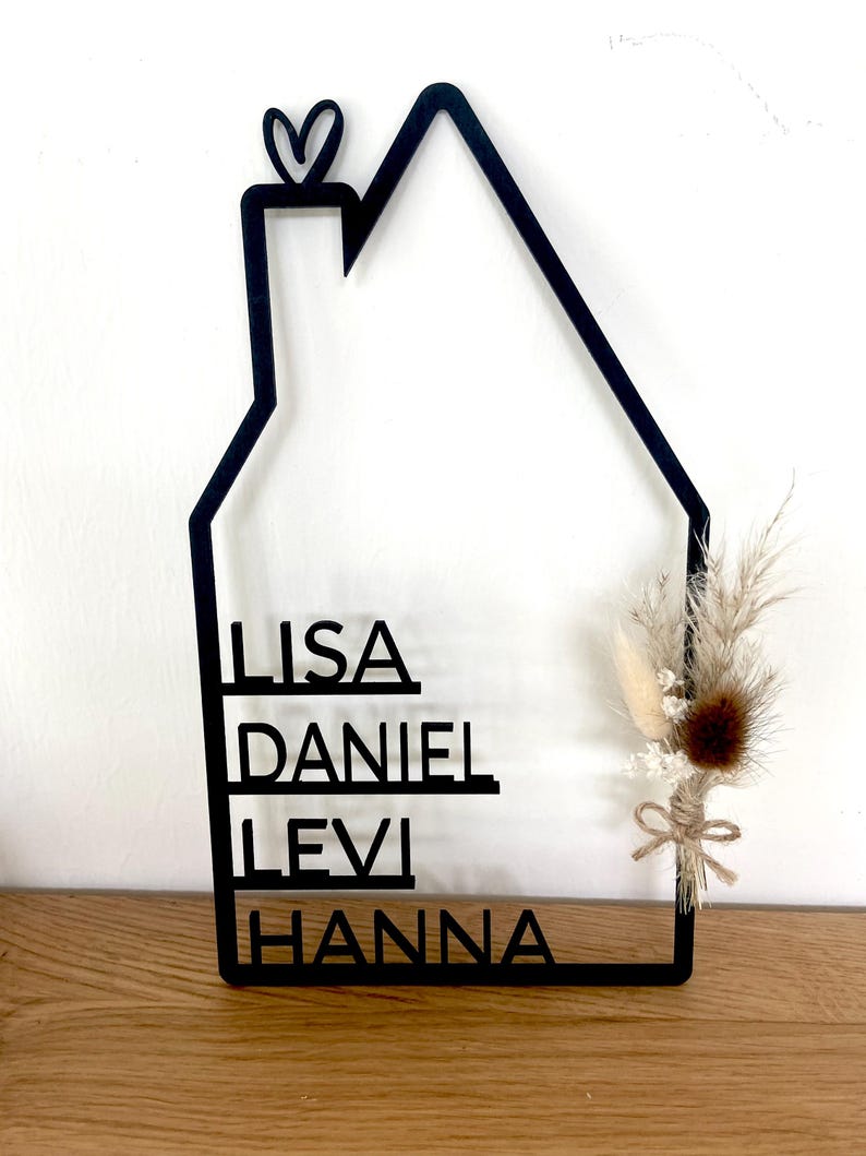 May include: Black wooden house-shaped sign with the names LISA, DANIEL, LEVI, and HANNA. A small heart is at the top. The sign is decorated with dried flowers and tied with twine. The sign is displayed on a wooden surface.