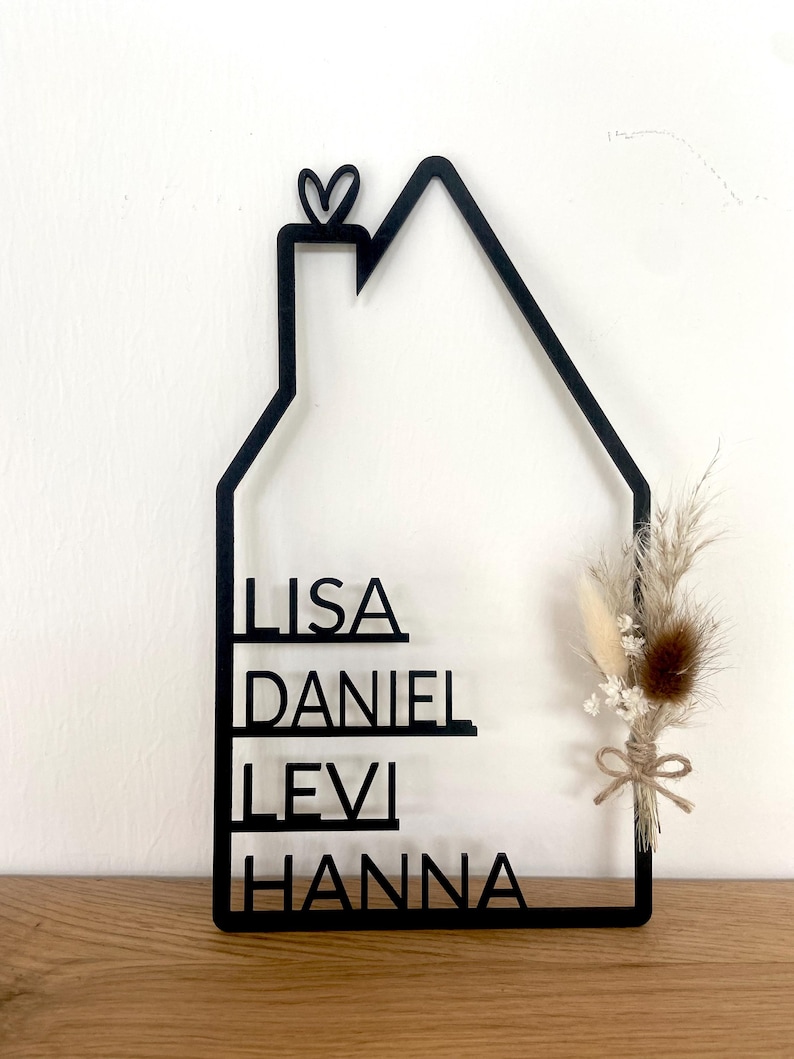 May include: A black, house-shaped wall hanging with the names LISA, DANIEL, LEVI, and HANNA cut out. A small heart tops the chimney. Dried flowers and a twine bow accent the right side. The piece is displayed on a wooden surface.