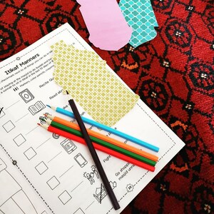 Op de afbeelding: Een zwart-wit werkblad met de titel "Itikaf Manners" en een lijst met activiteiten. Het werkblad ligt op een rood en zwart gestreept tapijt met kleurpotloden en papieren uitsnijdingen.