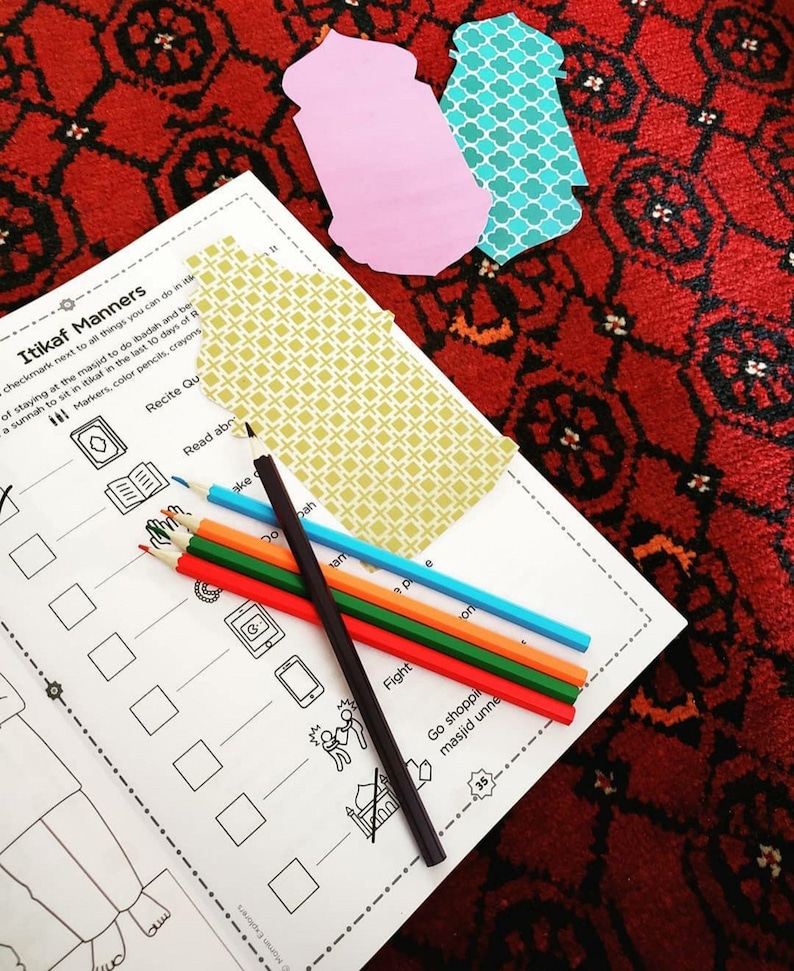 Op de afbeelding: Een zwart-wit werkblad met de titel "Itikaf Manners" en een lijst met activiteiten. Het werkblad ligt op een rood en zwart gestreept tapijt met kleurpotloden en papieren uitsnijdingen.