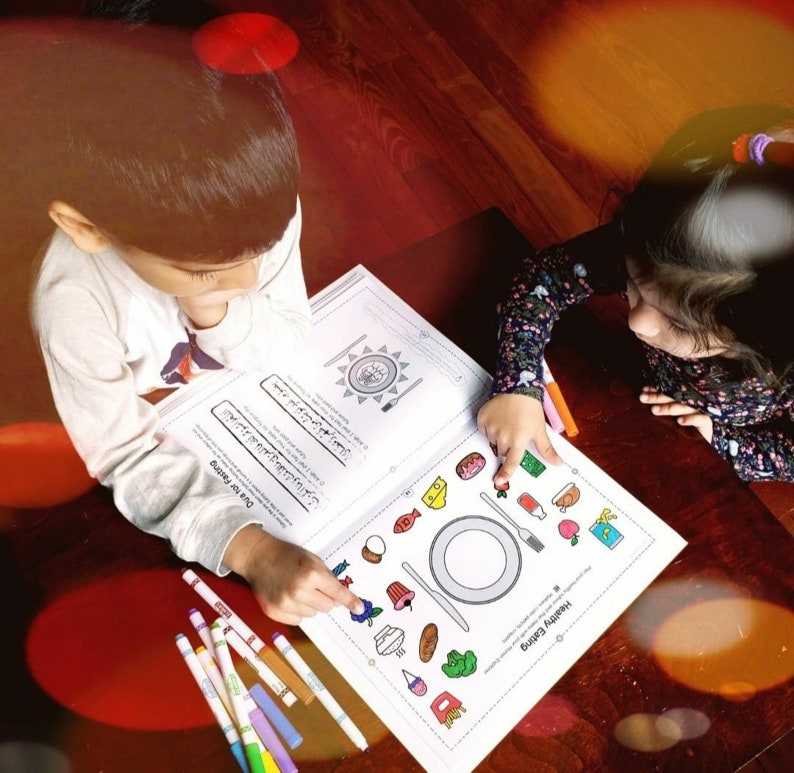 Op de afbeelding: Twee kinderen werken aan een kleurplaat met een afbeelding van een bord met gezond eten. De pagina heeft de titel "Gezond eten" en de tekst "Hou je van eten?" De kinderen gebruiken kleurpotloden om de afbeelding in te kleuren.