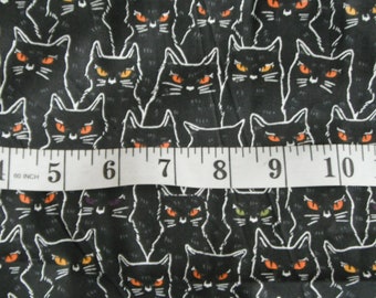May include: Black fabric with a repeating pattern of black cats with orange eyes. A white measuring tape with black numbers is placed across the fabric. The numbers on the tape read 3 to 12 inches.