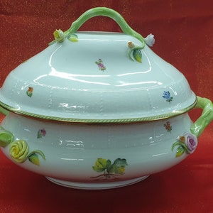 Puede incluir: Una fuente de servir de porcelana blanca con tapa y asas verdes. La fuente está decorada con diseños florales y un borde dorado.