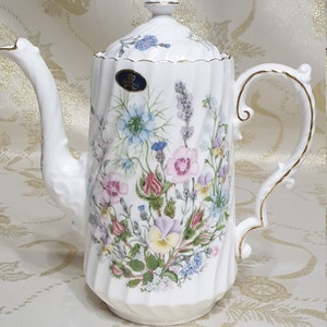 Könnte beinhalten: Weiße Porzellan-Kaffeekanne mit goldener Verzierung und Blumenmuster. Die Kanne hat einen Deckel mit einem Knopf oben und einen Griff an der Seite. Das Blumenmuster zeigt rosa, blaue und gelbe Blumen.