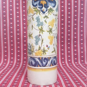 May include: A white ceramic vase with a yellow, blue and green floral pattern. The vase is cylindrical in shape and has a smooth, glossy finish.