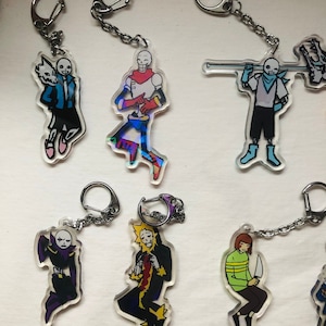 Peut inclure: Un ensemble de neuf porte-clés en acrylique colorés représentant des personnages du jeu vidéo Undertale. Les porte-clés sont attachés à des anneaux de clés argentés. Les personnages incluent Sans, Papyrus, Frisk, Chara et Mettaton.