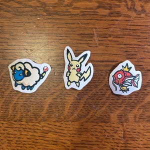 Puede incluir: Tres pegatinas troqueladas con personajes de dibujos animados. La primera es una oveja blanca esponjosa con cara azul y orejas amarillas. La segunda es una criatura amarilla con orejas largas y mejillas rojas. La tercera es un pez rojo.
