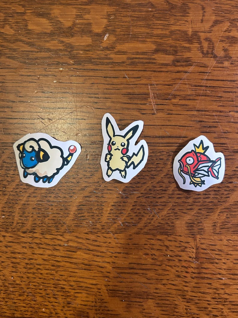 Puede incluir: Tres pegatinas troqueladas con personajes de dibujos animados. La primera es una oveja blanca esponjosa con cara azul y orejas amarillas. La segunda es una criatura amarilla con orejas largas y mejillas rojas. La tercera es un pez rojo.