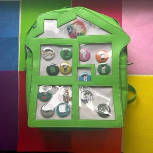 May include: A bright green backpack shaped like a house, with clear window panels displaying colorful pinback buttons. The backpack has a top handle and a zipper closure. The background features colorful squares.