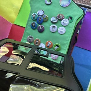 May include: A collection of colorful buttons and a keychain displayed on a green fabric. The buttons feature various cartoon character designs. A black bag with clear pockets is in the foreground, and a rainbow-colored background is visible.