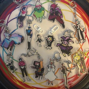 May include: A collection of acrylic keychains featuring various cartoon characters. The keychains have silver chains and clasps. The characters are in various poses and outfits, with a variety of colors. The keychains are arranged on a circular surface with a rainbow design.