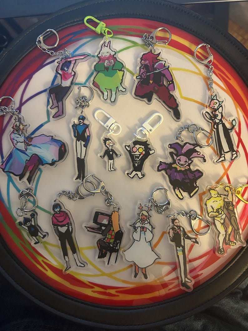 May include: A collection of acrylic keychains featuring various cartoon characters. The keychains have silver chains and clasps. The characters are in various poses and outfits, with a variety of colors. The keychains are arranged on a circular surface with a rainbow design.
