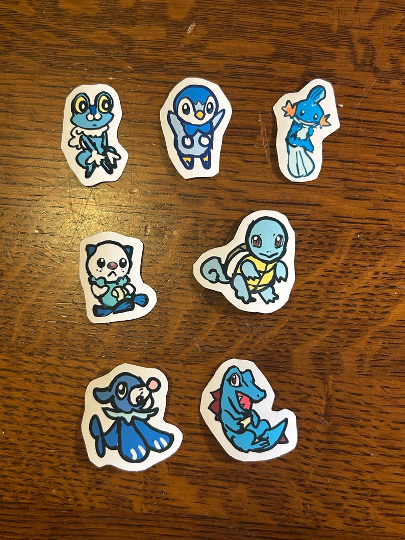 Puede incluir: Una colecci&oacute;n de siete pegatinas de papel individuales con ilustraciones dibujadas a mano de varias criaturas de dibujos animados en tonos azules. Cada pegatina tiene un borde blanco y representa un personaje &uacute;nico con expresiones faciales.