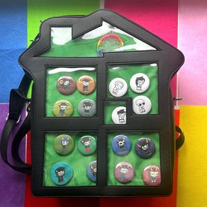 May include: Black house-shaped backpack with clear window panels displaying colorful character buttons. The backpack has a shoulder strap and a rainbow-colored decoration at the top. The background is a colorful patchwork of purple, blue, green, pink, and yellow.