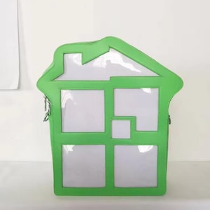 May include: A unique, house-shaped handbag with a bright green frame and clear, window-like panels. The bag features a zipper closure and a shoulder strap. The design is modern and playful, suitable for carrying essentials.
