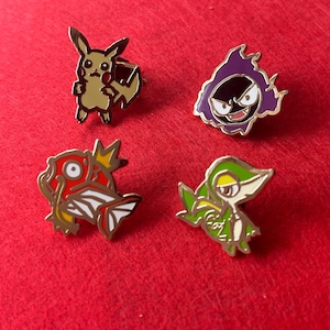 May include: Four enamel pins featuring cartoon characters. The pins include Pikachu, a purple ghost, a red and white fish, and a green and white snake-like creature. Each pin has a gold-colored border and is set against a red background.