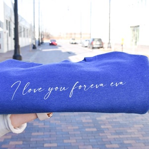 May include: A blue sweatshirt with the text "I love you forever eva" in a cursive font. The sweatshirt is held up in front of a blurred outdoor background.