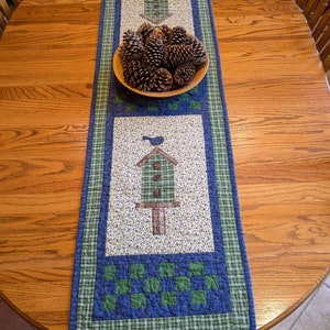 May include: A blue and green quilted table runner with a birdhouse design and pine cones in a bowl. The runner is on a wooden table.